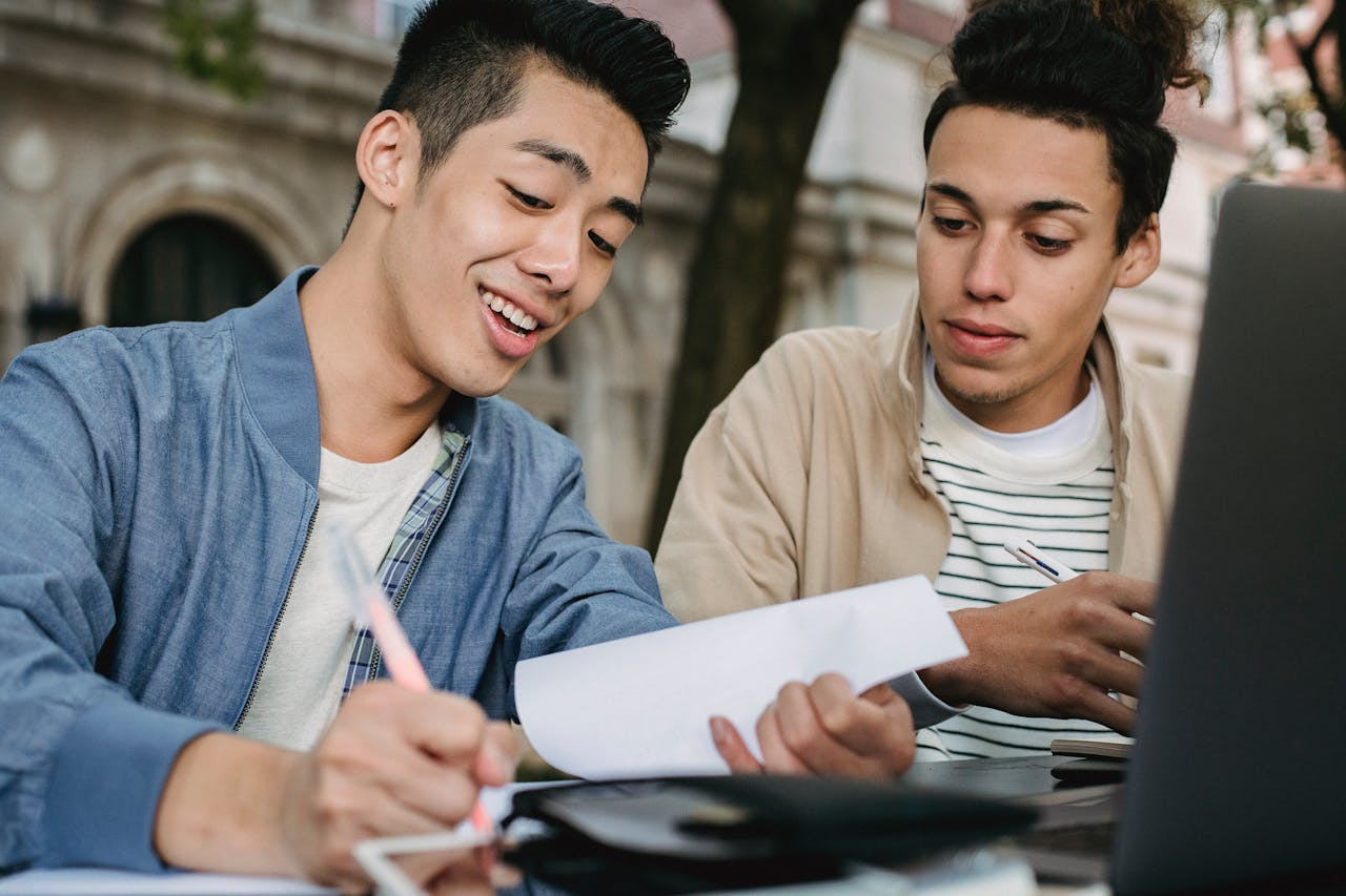Smiling ethnic male learner writing in document while doing homework assignment with classmate in park